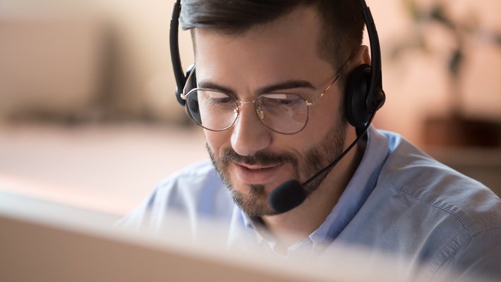 Nahaufnahme eines bärtigen Mannes mit Brille, der ein Headset trägt, auf einen Bildschirm blickt und mit jemandem telefoniert.